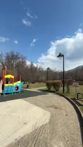 The camera pans right across a bright, sunny park.  A colorful, ship-shaped play structure sits on a rubberized pad to the left, near a concrete pathway that curves to the right. Along the path, there are park benches, short bushes, and lampposts.  Beyond the path is a grassy field with bare trees, leading to a mountain range in the background.  Clouds dot a bright blue sky.  There are structures resembling small cabins visible behind the trees. A tree stump stands prominently in the field. The day is clear and sunny.