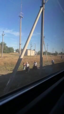 The video shows a bright, sunny day with a clear blue sky. The camera appears to be inside a vehicle, looking out a window. In the foreground, a dark, reflective window frame is visible. Beyond the window, a dry, grassy field stretches out, dotted with scattered trees and bushes. Several utility poles, some with diagonal support beams, are prominent features in the landscape. A tall, thin tower with a platform at the top stands in the background. A low, brick building is also visible in the distance. There are four people in the field. Three of them are standing near a utility pole, and one is further away, walking to the right. The three people near the pole appear to be older adults, two men and one woman. The woman is holding a bouquet of flowers. They are all dressed casually. The fourth person, a man, is walking away from the camera towards the right side of the frame. The overall impression is of a rural or semi-rural setting, possibly a roadside or a field near a small settlement.