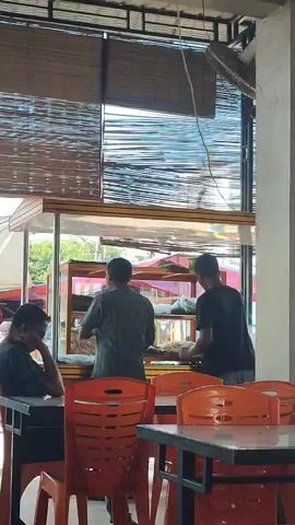 The video shows a man in a green shirt standing behind a display case filled with food. He appears to be serving a customer. To his right, another man in a black shirt is also behind the counter, engaged in a similar activity. In the foreground, a man in a dark shirt sits at a table, facing away from the counter, his head tilted. The environment appears to be a casual dining establishment, possibly an outdoor eatery or a place with open-air seating, indicated by the bamboo blinds and the bright natural light filtering in. The tables are set with orange chairs.