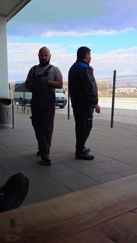 Two men are standing outside on a tiled patio. The man closest to the camera is wearing gray overalls over a gray t-shirt and black shoes. He has a beard and is looking at the camera and giving a thumbs up. The other man is standing behind him and to his right. He is wearing a dark jacket with blue accents, dark pants, and dark shoes. He is smoking a cigarette and looking off to the right. There is a metal trash can to the left of the man closest to the camera. Behind them is a wire fence, a white van, and a few chairs. The sky is blue with some clouds. The video is shot from a low angle, and a pair of dark shoes can be seen in the bottom left corner of the frame.