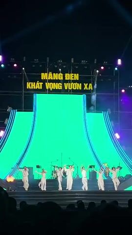 The video captures a live stage performance featuring seven individuals dressed in white outfits. They are positioned on a black stage with a large backdrop displaying the words "MANG DEN KHAT VONG VUON XA". The lighting on stage includes purple and blue spotlights, and there's an animated light display on the stage itself. The performers are engaged in a dance routine, with one of them holding a microphone. In the foreground, a crowd of people is visible, watching the performance. The scene is dynamic and energetic, typical of a live music or dance show.