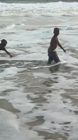 The video shows a beach scene. Three people are visible. Two are children, and one is an adult. The children are in the water. One is crawling in the surf, while the other is walking and playing with a white disc, possibly a frisbee. The adult is standing on the beach, partially blocking the view. The sea is choppy, with white foam visible on the waves. The sky is overcast.