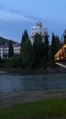 The scene is an outdoor shot of a river with buildings in the background. The river flows from right to left. On the right side of the river, there's a bridge with lights. On the left side, a white building with a tower topped with a crown can be seen. Next to it is another white building with red roofs. Tall trees are interspersed between the buildings. The sky is overcast, suggesting it's either early morning or late evening. The landscape appears to be mountainous, with a hill visible in the background. There is green grass in the foreground.
