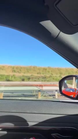 The video shows the interior of a car looking out the window. The car is on a highway with green hills in the background and a bright blue sky. A red sports car is visible in the side mirror and ahead of the car. A smiley face air freshener with sunglasses is hanging from the rearview mirror. The steering wheel and dashboard of the car are also visible. The video appears to be filmed while driving on a sunny day.