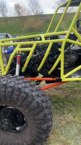 The video shows a yellow, open-frame buggy, likely homemade, with a black engine and transmission visible through the frame. It has two seats, big treaded tires, and a battery visible in the rear.  The frame has orange supports.  The buggy is parked on a grassy field. In the background is a nondescript landscape, a light-colored car, and other blurred vehicles. Music plays loudly.