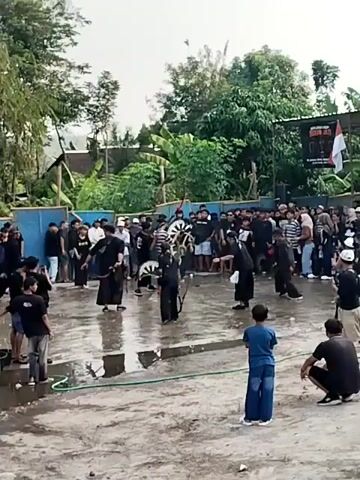 This media file captures a vibrant outdoor gathering in Gentungan, Indonesia. The scene is filled with a crowd of people, many of whom are dressed in black attire, suggesting a unified group or performance. At the center of the activity, a group of individuals in black clothing are engaged in what appears to be a dynamic performance. Some are holding what look like traditional instruments or props, possibly a type of drum or elaborate headdress. The ground is wet, indicating recent rain or a deliberate watering, creating reflections that add to the visual texture of the scene.

The setting is an open area, possibly a street or courtyard, with buildings and lush green trees forming the backdrop. A banner with some text is visible in the upper right corner, partially obscured by trees. A prominent Indonesian flag is also displayed, hinting at a national or cultural event. The atmosphere appears lively, with many spectators observing the performance. The presence of children in the foreground suggests a family-friendly event. The overall lighting suggests it is daytime, with overcast skies. The activity seems to be a cultural performance or parade, drawing a significant local audience.