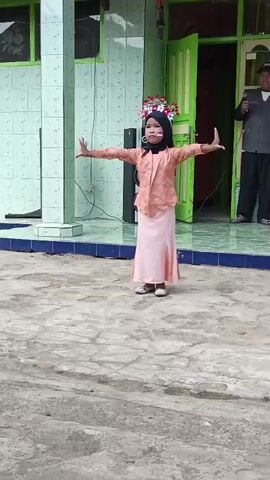 A young girl wearing a light peach and pink outfit and a black hijab is performing a dance in an outdoor area. She has flowers in her hair and red and white face paint on her cheeks. She is standing on a concrete surface in front of a building with green and white tiles and a green door. There are two speakers behind her. To the right of the frame, an adult male wearing a dark vest over a white shirt and white pants is holding a microphone and appears to be speaking or singing. Another person in a red vest is visible behind him. The area is somewhat cluttered with wires and debris on the ground.