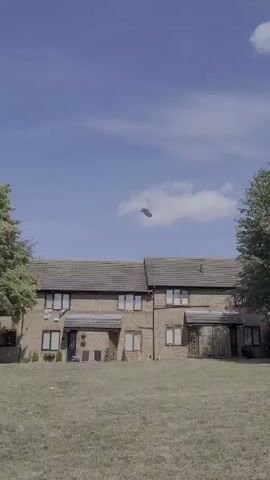 A drone is visible flying in the sky above a row of houses. The houses are made of brick and have tiled roofs. There is a patch of dry grass in front of the houses, with trees on either side. The sky is blue with some clouds.