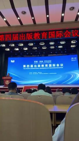 The video shows a conference hall with a large screen displaying "THE FOURTH INTERNATIONAL CONFERENCE ON PUBLISHING EDUCATION". A speaker stands at a podium on the left side of the stage, addressing an audience seated in rows of chairs. The audience members are facing away from the camera, their backs visible. The hall is illuminated by overhead lights and spotlights focused on the stage. The overall atmosphere appears to be formal, consistent with an academic or professional conference.