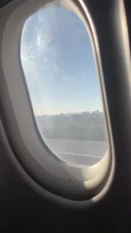 The video shows a view through an airplane window. Outside, the sky is blue with some scattered clouds. Below, a cityscape is visible in the distance. The ground appears to be a runway or tarmac, with some buildings and vegetation. The lighting suggests it is daytime, possibly morning or late afternoon. The perspective is from inside the cabin, looking outwards.