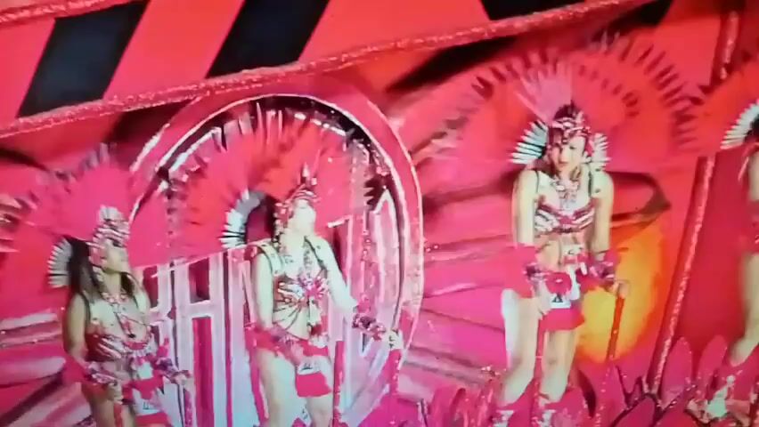 The video shows a performance with four women dressed in elaborate, feathered costumes with headdresses. The costumes are predominantly red and pink, with metallic accents. They are dancing on a platform with a bright pink, glittery background and large circular cutouts. The women are positioned in a line, and their movements are synchronized. One woman on the right appears to be holding a staff or cane. In the background, there are black and red diagonal stripes. The lighting is bright and appears to be stage lighting. The video appears to be a recording of a live event, possibly a parade or a stage show.