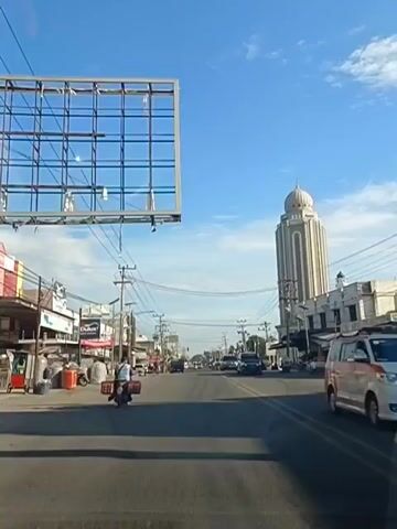 The video shows a daytime view of a street with a wide paved road. On the left side of the road, there are buildings, some with advertisements and goods displayed outside. A billboard is visible in the upper left corner of the frame. Several vehicles, including cars and motorcycles, are on the road, and some people are riding on motorcycles, one pulling a cart. On the right side of the road, there is a distinctive tall, light-colored building with a dome at the top, resembling a minaret or a tower, and other buildings. The sky is blue with scattered clouds, and power lines are visible crisscrossing above the street. The camera appears to be moving forward, suggesting it is from a vehicle.