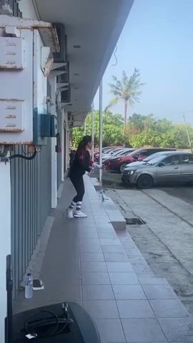 A woman is standing on a tiled walkway next to a building with metal siding and air conditioning units. She is wearing athletic clothing and appears to be dancing. She is holding a bottle in her right hand. There are parked cars in the background, including a silver hatchback and several red cars. A palm tree is visible in the distance. Another person is standing near the cars on the right side of the frame. There is a white bottle and a smartphone on the walkway near the woman.