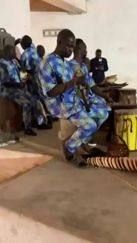 A group of five men are playing traditional African musical instruments indoors, accompanied by drumming.  The men are wearing matching blue and green patterned outfits.  They are seated on stools or benches.  One man is holding and shaking a gourd-like instrument with a long handle. Another man is shaking a smaller, round instrument. A third man is playing a drum.  There is a second drum and what looks like a percussion instrument made of stacked wooden disks on the floor in front of them.  Another man in a black suit is standing and appears to be observing.  The setting appears to be a bare room with simple decor, with beige walls and floor.  There are some bags and other objects in the background.
