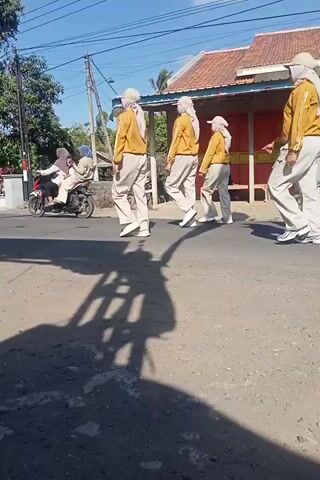 A group of people in matching yellow jackets and white pants are standing in a line on a paved road, facing to the left. The video is shot from a low angle, looking up at the people. The lighting suggests it is daytime, with shadows visible on the road. There are buildings and trees in the background. The people appear to be performing some sort of synchronized movement or drill.