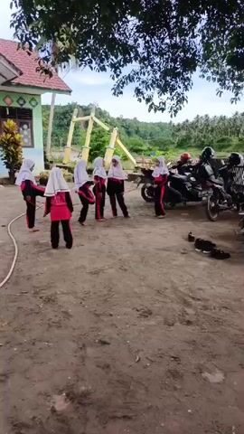 A group of six young girls, all wearing matching red and black tracksuits and white headscarves, are gathered in a dirt courtyard. They appear to be in a schoolyard setting. One girl holds a long white hose on the ground. They stand relatively still, seemingly preparing for an activity. Behind them, two motorcycles are parked next to a pair of shoes. In the background, there is a green building with a red roof and a yellow decorative plant, along with a yellow, A-frame structure. A tree with green leaves overhangs the scene from the top. The sky is overcast, and there is a view of trees and a hillside in the distance.