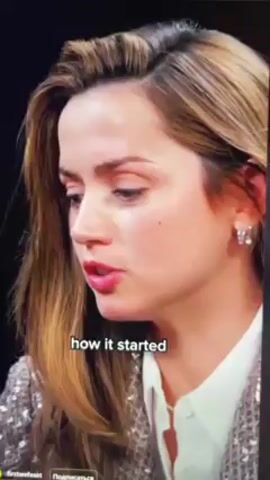 Here is a description of the video:

A close-up shot shows actress Ana de Armas. She is speaking and the caption "how it started" is on the screen. She is wearing a white button-down shirt and a sequined blazer. Her hair is blonde, and she is wearing small earrings. She looks surprised when she hears a sound off screen and touches her hair.