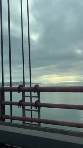 A video shows a view from a bridge overlooking a body of water. A red pole with a warning sign featuring a triangle and curved lines is visible in the foreground on the left. The sign appears to depict wind or currents. Further along the pole, the numbers "1" and "4" are affixed. Red railings run horizontally across the middle of the frame. The water is calm and reflects the overcast sky, with several ships visible in the distance. The coastline and a city skyline are discernible on the left horizon. The overall atmosphere is muted due to the cloudy weather.