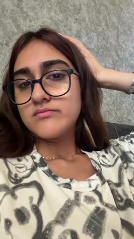 A young woman with brown hair and glasses makes a kissing face and gestures with her hands in front of her face. She is wearing a white t-shirt with a black and white pattern. The background appears to be a textured wall.