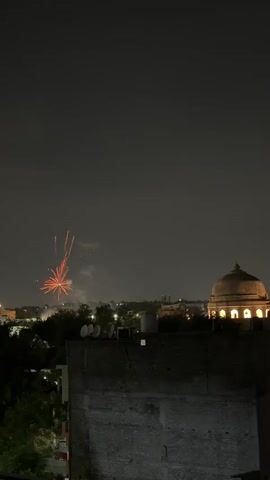The video shows a nighttime cityscape with fireworks in the distance. A dome-shaped building is visible on the right side of the frame, illuminated by lights. In the foreground, the top of a dark, brick building is visible, with some satellite dishes on its roof. The fireworks are colorful bursts of light against the dark sky, with some smoke visible. The fireworks appear to be exploding at various heights and locations across the horizon.