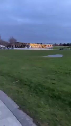 The video starts with a wide shot of a grassy area with a small puddle. In the background, there is a building-like structure with lights. The camera then rapidly pans across the grassy area, revealing a paved walkway. The video concludes with a closer view of two massive, silver horse head sculptures, constructed from individual metal pieces. The sculptures stand on a grassy mound, set against a cloudy sky.