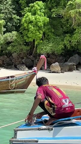 A man wearing a maroon and yellow shirt with the number 71 on the back is crouching on a boat, holding a rope. He is in the process of tying or untying the rope attached to another boat. The other boat is a longtail boat with wooden accents and decorations. In the background, there is a sandy beach and lush green trees on a rocky hillside. The water is clear and turquoise.