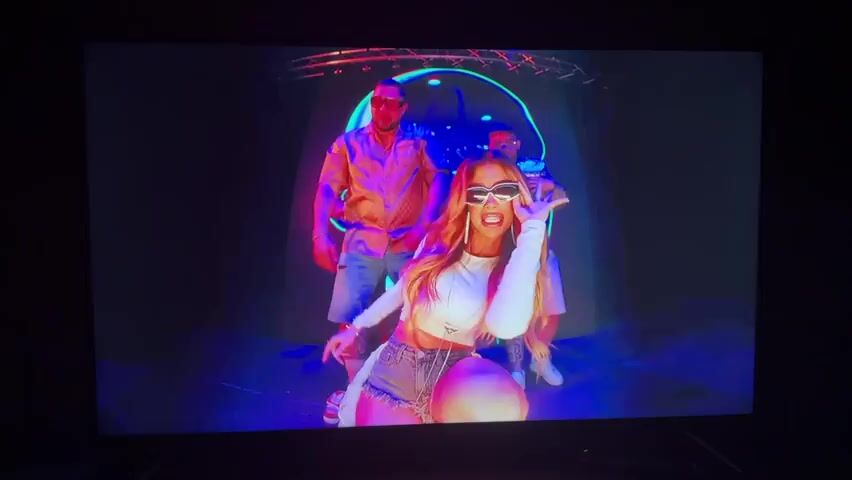 The video opens with a blurry, colorful shot of people dancing in what appears to be a nightclub or stage setting with neon lights. The lighting is predominantly blue and pink. The scene transitions to a man in a white t-shirt and denim shorts standing in a red room with neon lights. A motorcycle is parked behind him, and there are some metallic objects and a small structure resembling a house. The man gestures with his hands. The scene then cuts back to the colorful club setting with two people dancing and posing. The man on the right is wearing sunglasses and a patterned shirt. The woman on the left is wearing a white top and denim shorts. The video ends with a close-up of a man wearing oversized orange-tinted sunglasses and holding a championship belt. He is speaking directly to the camera.