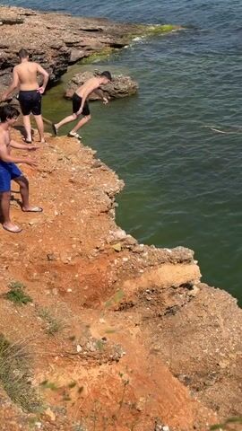 The video captures a scenic coastal setting in Νέα Χηλή, Greece, on a clear, sunny day. A young man, wearing black shorts, is shown in mid-air having just jumped from a reddish-brown rocky cliff into the blue-green sea. The camera briefly shows him splashing into the water. Another young man, also in black shorts, stands near the cliff edge, appearing to watch or prepare for his own jump. A third person's legs and feet, wearing flip-flops, are partially visible on the left side of the frame. The sea is calm with gentle waves, and distant land or mountains are visible on the horizon under a clear sky. There is no visible text in the video.