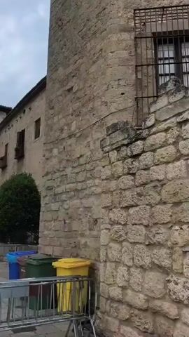 The video begins by showing a stone wall with a window at the top. The scene then shifts to reveal a large stone building with a grand entrance featuring a rose window above it. Several people are walking around in the courtyard in front of the building. Two women are walking together towards the entrance. Further into the courtyard, a couple is walking hand-in-hand. Some people are standing and looking at the building. The sky is cloudy.