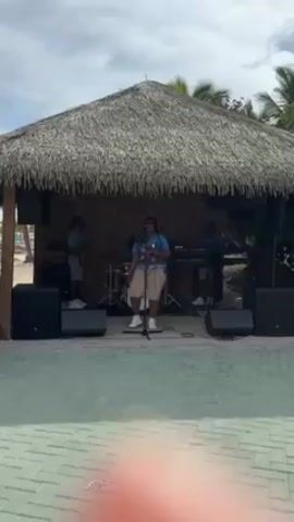 A medium shot captures a band performing on an outdoor stage beneath a thatched roof structure. The singer, a person with dark skin wearing a patterned blue shirt and khaki shorts, stands at the microphone in the center of the stage. Behind them are the other band members with their instruments: drums and a keyboard. Large black speakers flank the stage on both sides. The flooring in front of the stage is composed of green and white tiles. In the background, there is a sandy area that appears to be a beach under a partly cloudy sky. A finger appears momentarily in the bottom center of the screen.