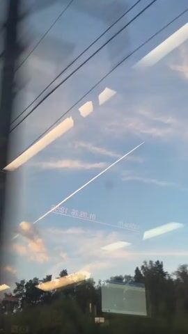 The video is a first-person view from a moving vehicle, likely a train, looking out a window. Overhead, several parallel dark wires are visible against a blue sky with scattered white and pink clouds. A long, thin white contrail from an airplane stretches diagonally across the sky. Reflections on the window obscure parts of the view, showing indistinct shapes and text. The bottom of the frame shows a blurry line of trees and foliage, with a rectangular screen displaying text, possibly an advertisement or information board, partially visible. The overall impression is a fleeting glimpse of a journey through a landscape under a dynamic sky.