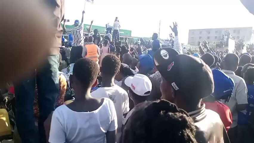 A large crowd of people, primarily adults, is gathered outdoors in what appears to be a street or open urban area in Mwingi, Kenya. The scene is brightly lit by the sun, suggesting daytime, possibly late afternoon. Many individuals have their hands raised in the air, indicating excitement, cheering, or celebration. People are closely packed together, with some standing on elevated structures or vehicles to gain a better view.

The crowd is diverse in attire, with various colors of shirts and caps visible, including blue, white, and black hats, and shirts in white, blue, and striped patterns. One person is seen wearing an orange vest. Buildings are visible in the background, further confirming an urban setting. A green structure with a partially visible "M-PESA" sign can be seen behind the crowd, a common mobile money service in Kenya, providing a strong location cue. Other visible text includes "NEW" on a black cap and "DEN" partially visible on a blue cap or jersey. The overall atmosphere appears lively and energetic, filled with collective enthusiasm.