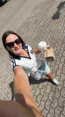 A woman with long dark hair and sunglasses stands in a parking lot, holding a McDonald's bag and a large white McDonald's cup. She is wearing a white v-neck shirt with blue stripes, denim shorts, and light blue shoes. She smiles and turns the camera slightly. The parking lot is paved with patterned bricks. A car is parked in the background, and there are trees visible.
