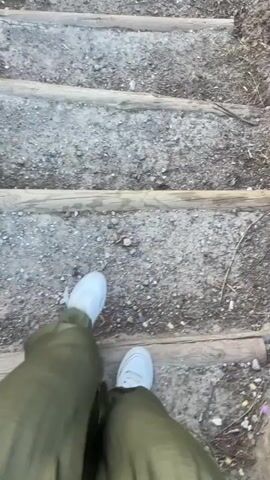 The video shows a first-person perspective of someone walking down a set of wooden steps. The steps appear to be made of rough logs embedded in a dirt or gravel path. The person is wearing white sneakers and green pants. The environment seems to be outdoors, possibly a trail or park, with some dry vegetation visible in the background. The camera is shaky, indicating movement.