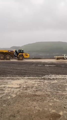 The video shows a large yellow dump truck driving on a muddy surface in a quarry on a cloudy day. The truck is seen from the side, then through a truck mirror showing multiple trucks behind it, and finally from inside the cabin looking out over the quarry and a large pile of gravel. The surrounding landscape includes hills and visible areas where earth has been removed, indicating mining or excavation operations. The ground is wet and muddy, suggesting recent rain.