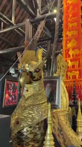 The camera pans up to show the interior of a large, wooden building. In the foreground is a golden, bird-like statue with intricate carvings, holding a framed portrait. Behind the statue, a tall, red banner hangs, adorned with golden animal figures. The building's wooden support structure is visible, with carved details and hanging banners in the background. The lighting is dim, with some bright spots.