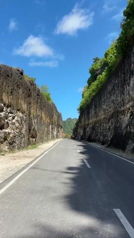 The video shows a straight, paved road running through a narrow passage formed by tall rock walls on either side. The walls are textured and vary in color, with some areas showing signs of vegetation. The road has painted white lines, and in the distance, several people are gathered near an orange structure. The sky above is blue with scattered white clouds. The overall environment appears to be sunny and outdoors.