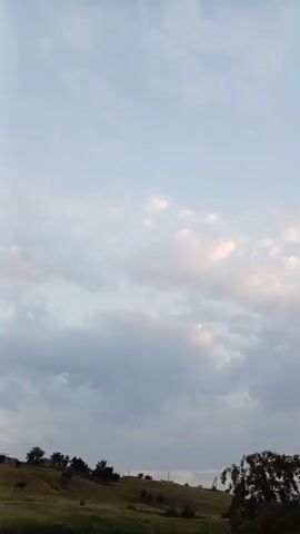 A video pans across a cloudy sky with soft pink and white hues, possibly at sunset or sunrise. Below the sky, a hilly landscape is visible with green grass, trees, and a small structure with a white roof. The overall scene suggests an outdoor, rural environment.