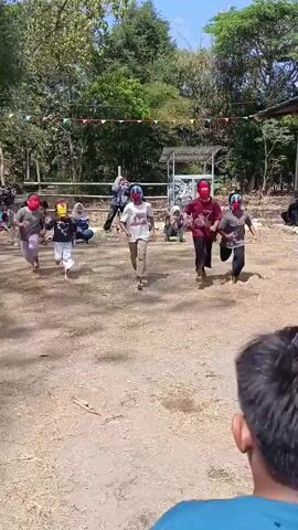 A group of children wearing masks are participating in an outdoor activity. They are lined up in two rows facing each other. The children in the front row are bent over, reaching into a black bucket. The children in the back row are also bent over, appearing to be assisting or participating in the same activity. Some of the children are wearing red masks, one is wearing a yellow and red mask, and another is wearing a blue and red mask. They are in an outdoor area with dry ground and trees in the background. There are spectators watching the activity.