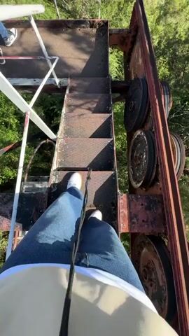 The video shows a first-person view of someone descending a set of metal stairs attached to a rusty metal structure with large wheels. The person is wearing jeans and white shoes. As they descend, the view opens up to reveal a lush green valley with mountains in the distance, and a glimpse of the ocean under a clear blue sky. In the valley, a tall red metal tower stands amidst the trees.