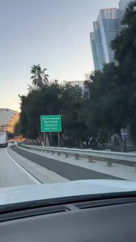 The video shows a view from inside a car driving on a highway. A green sign reading "Hollywood Burbank Airport NEXT RIGHT" is visible. Trees and buildings line the side of the highway. The sky is clear and appears to be dusk. There is other traffic on the highway, including trucks and cars. The camera is focused on the road ahead and the scenery passing by.