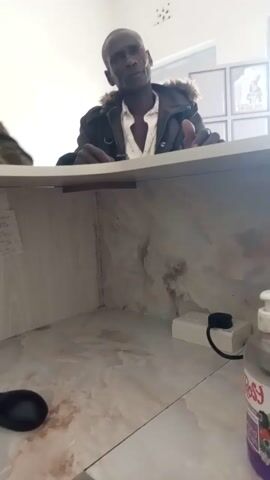 A low-angle, slightly blurry video shows a man in a jacket and open shirt standing behind a desk. The desk surface appears to be made of a light-colored material with a marble-like pattern. There are some stains on the surface. On the desk, there is a black computer mouse and a black keyboard. A black electrical cord runs from the keyboard and disappears out of frame. In the background, there is a white electrical outlet with a black plug connected to it. Further in the background, there is a white wall with some framed items. Several small pieces of paper are attached to the wall of the desk cubby. The man is looking at the camera and gesturing with his right hand. The video is shot from a perspective that is looking up at the man and the desk.