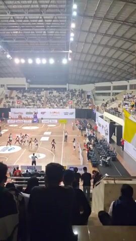 The video shows a basketball game taking place in an indoor arena. The court is visible with players on it, along with spectators in the stands. The arena appears to be quite large with a high ceiling. The lighting is typical for an indoor sports venue. The camera angle is from the perspective of someone seated in the stands, looking down at the court. The video is shaky, suggesting it was filmed by hand. The focus shifts periodically, but the overall scene remains consistent throughout the short clip. There are advertisements visible around the court, indicating sponsorship of the event. The spectators are diverse in appearance and clothing. The actions on the court suggest an active game is in progress, though specific plays are not clear due to the distance and camera movement. The overall atmosphere seems to be one of a live sporting event.