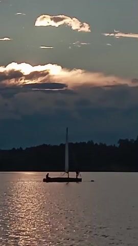 The video shows a sailboat on a body of water during sunset. The sun is partially obscured by clouds, casting a warm glow on the water. The sailboat has two people on it, one sitting and one standing. The background is a silhouette of trees against the colorful sky. The camera is panning slightly to the right.