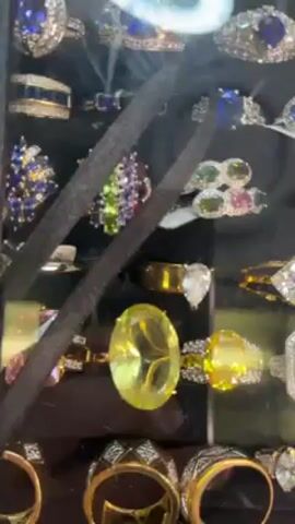 The video shows a display case full of rings. The rings are a variety of colors, shapes, and sizes, with some set in gold or silver colored metal. The rings are lit from above.