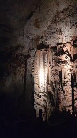 The video shows a large, illuminated rock formation in a cave. The formation is composed of stalactites and stalagmites, creating a textured and layered appearance. The rock is primarily a light brown or beige color, with some reddish-brown hues. A spotlight is directed at the center of the formation, highlighting its intricate details and casting shadows. The surrounding area of the cave is dark, with only parts of the ceiling and walls visible. The overall impression is one of natural grandeur and geological artistry.