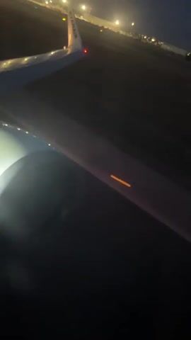 The video shows a view from an airplane window at night. The wing of the plane is visible in the foreground, with the Ryanair logo on the wingtip. The plane is on a runway, indicated by the lights in the distance. The engine is also partially visible, illuminated by its own light. The runway is dark, with scattered lights visible along its edge and in the distance, suggesting a city or airport environment. The plane appears to be moving, as the lights are streaking, and there is a slight blur to the image. The overall impression is of a nighttime takeoff or landing.