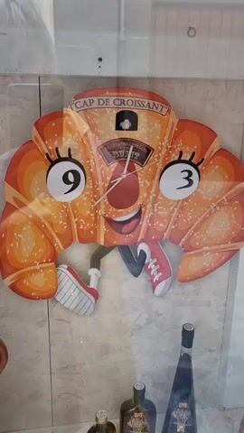 A cartoon clock shaped like a croissant with a face hangs on a wall. The clock has hands pointing to approximately 9:15. The clock face is orange with sesame seeds and has cartoon eyes and a smiling mouth. It has arms and legs with shoes. Below the clock are several bottles, one of which is a dark glass bottle with a tall neck. The background appears to be a marble-like tiled wall.