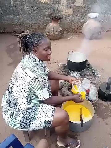 A woman in a white and black patterned short-sleeved shirt and shorts is squatting on a small wooden stool, adding liquid from a plastic bag to a large pot of yellow liquid. She stirs the pot with a large metal utensil. A fire burns nearby, with a pot on top of it, emitting steam. Behind her, there are earthen pots and a brick wall. A metal kettle sits near the fire. A small blue chair is visible to the left, with someone sitting in it. Another person walks in the background near a corrugated metal structure. The ground is dirt.