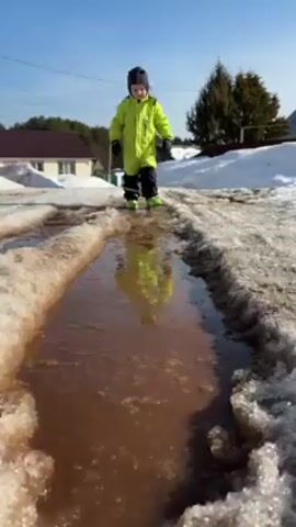 A young child is walking through a muddy puddle. The child is wearing a bright green snowsuit with black gloves and black boots. They also have a dark gray knit cap on their head. The puddle is long and narrow, situated on a path with snow on either side. The water is murky brown, reflecting the child's figure. In the background, there's a house with a brown roof, some trees, and a clear blue sky.